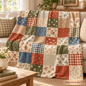 May include: A patchwork throw blanket featuring a grid of square fabric panels with diverse patterns including red and white gingham, blue and white stripes, and various floral prints in shades of red, green, blue, and cream on a textured fabric.