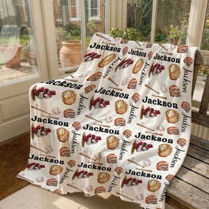 Op de afbeelding: Een witte baseball-thema deken met een herhalend patroon van honkballen, handschoenen, helmen en knuppels. De naam "Jackson" staat herhaaldelijk in het zwart op de deken gedrukt. De deken is over een houten bank gedrapeerd.