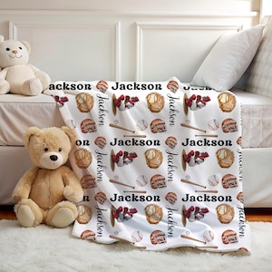 Op de afbeelding: Witte baseball-thema deken met herhaalde patronen van honkbalhandschoenen, helmen, honkballen en knuppels. De naam "Jackson" is geschreven in een scriptlettertype. De deken ligt op een bed met twee teddyberen.