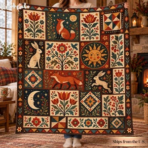 May include: A rectangular throw blanket featuring a grid of folk-art style illustrations including foxes, rabbits, a sun, a moon, and floral motifs in a palette of deep red, navy blue, cream, and orange, with a decorative geometric border.