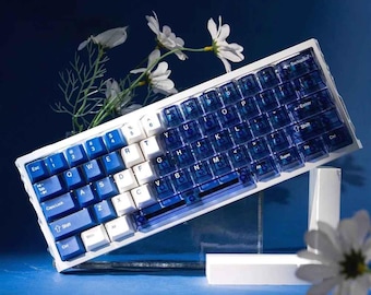 May include: A mechanical keyboard with a white frame and blue and white keycaps. The keycaps are a mix of solid white and translucent blue. The keyboard is angled, with a few white flowers in the background. The keyboard has a standard QWERTY layout.