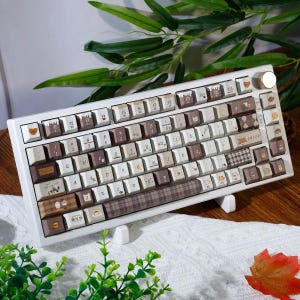 Puede incluir: Un teclado mecánico blanco con teclas personalizadas en tonos marrón, crema y beige. Las teclas muestran varios patrones, incluyendo cuadros, ilustraciones relacionadas con el café e iconos pequeños. Un mando metálico circular se encuentra en la parte superior derecha.