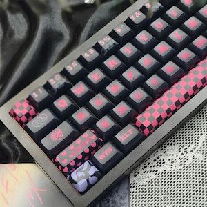 May include: A black mechanical keyboard with pink lettering and accents. The keys are black with pink letters, and some keys feature heart and checkerboard patterns. The keyboard has a dark grey frame.