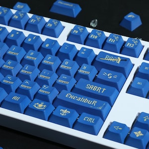 Könnte beinhalten: Eine mechanische Tastatur mit blauen Tastenkappen und einem weißen Rahmen. Die Tastenkappen haben goldfarbene Schrift und Symbole, darunter das Wort "Excalibur". Zusätzliche Tastenkappen liegen um die Tastatur herum.