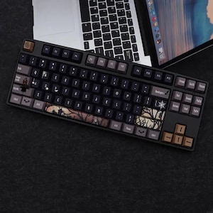Puede incluir: Un teclado mecánico negro con teclas personalizadas con temática de Halloween. Las teclas tienen una combinación de colores oscuros con acentos marrones y grises. El teclado está colocado sobre una superficie oscura, con un portátil en el fondo.