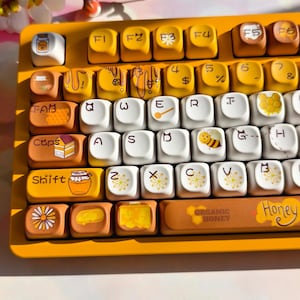 May include: A custom mechanical keyboard with yellow and white keycaps. The keycaps feature honey-themed designs, including bees, honeycombs, and jars of honey. The keyboard has a yellow frame and the word "Honey" is printed on the spacebar.
