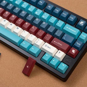 Könnte beinhalten: Eine schwarze mechanische Tastatur mit Tasten in Blau-, Weiß- und Burgundertönen. Die Tastatur hat eine Vielzahl von Funktionstasten und eine separate burgunderfarbene Taste mit einem weißen Pluszeichen. Die Tasten sind in einem Standard-QWERTZ-Layout angeordnet.