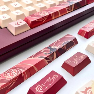 May include: Close-up of a mechanical keyboard with custom keycaps. The keycaps feature a red and cream color scheme with intricate designs and Japanese characters. The keyboard's frame is a deep red color.