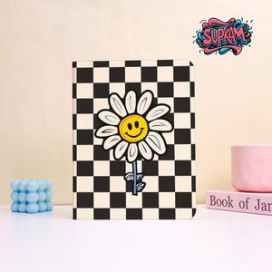 May include: A notebook with a black and white checkered pattern and a large daisy graphic. The daisy has a yellow center with a smiling face and white petals. The notebook has the word "SUPKAM" in a colorful graffiti style.