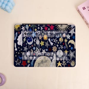 Peut inclure: Une coque d'ordinateur portable bleu fonc&eacute; avec un design c&eacute;leste color&eacute;. La coque est orn&eacute;e d'illustrations d'&eacute;toiles, de plan&egrave;tes, de lunes et de soleils de diff&eacute;rentes couleurs. Du texte blanc est imprim&eacute; au centre de la coque.