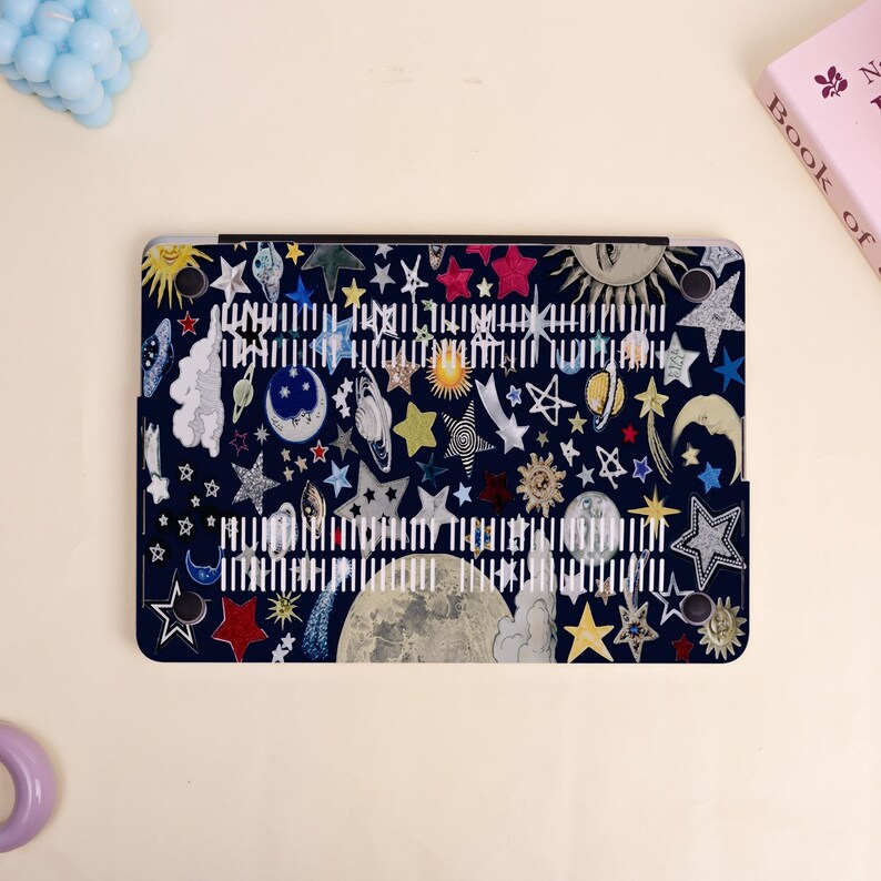 Peut inclure: Une coque d'ordinateur portable bleu fonc&eacute; avec un design c&eacute;leste color&eacute;. La coque est orn&eacute;e d'illustrations d'&eacute;toiles, de plan&egrave;tes, de lunes et de soleils de diff&eacute;rentes couleurs. Du texte blanc est imprim&eacute; au centre de la coque.