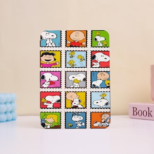 Può includere: Una custodia rettangolare con un collage di personaggi dei Peanuts in disegni di francobolli. I francobolli mostrano Snoopy, Charlie Brown e altri personaggi in varie pose, su uno sfondo bianco con un bordo colorato.