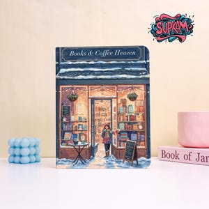 May include: A rectangular notebook featuring a winter scene of a bookstore storefront with the text Books & Coffee Heaven. The illustration shows a person in a scarf standing in the doorway, surrounded by snow, bookshelves, and warm string lights.