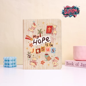 Peut inclure: Un journal en bois avec le texte "My Hope is in Jesus" en lettres colorées. La couverture présente des illustrations d'agneaux, d'une croix, de fleurs et d'un palmier. Le journal a un motif de grain de bois clair.