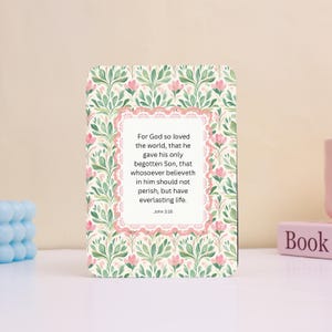 May include: A floral-patterned card with a quote from John 3:16. The card features a cream background with green leaves and pink flowers. The quote is in a white frame with a pink lace border. A small pink book is visible.