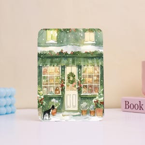 以下が含まれることがあります： クリスマスの飾り付けがされたお店の水彩イラスト。お店には緑色のオーニング、白いドア、プレゼントでいっぱいの窓があります。お店の前には白黒の犬が立っています。