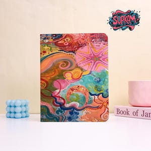 May include: A rectangular notebook featuring a vibrant, abstract design with swirling patterns in shades of pink, orange, blue, and yellow. The cover displays a prominent star-like shape and organic curves. The text SUPKAM is visible in the top right corner.
