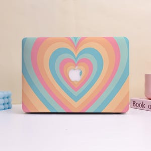 Peut inclure: Une coque d'ordinateur portable avec un motif de cœur coloré. L'étui présente un motif de cœur répété dans des tons orange, rose, bleu et vert. Le logo Apple est visible au centre du motif en forme de cœur.