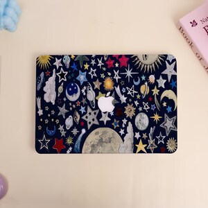 Peut inclure: Une peau d'ordinateur portable bleu marine avec un motif c&eacute;leste. Elle pr&eacute;sente des &eacute;toiles, des lunes, des plan&egrave;tes et des soleils de diff&eacute;rentes couleurs. Le logo Apple est blanc. Motif fantaisiste sur le th&egrave;me de l'espace.