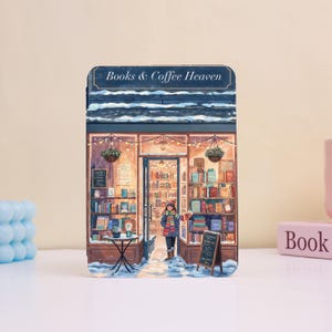 May include: A decorative item depicting a bookstore scene with the text "Books & Coffee Heaven." The illustration shows a person in the doorway, surrounded by bookshelves, snow, and hanging flower baskets, creating a warm atmosphere.