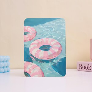 May include: A rectangular art print featuring a painting of three pink and white striped inflatable rings floating in a pool of blue water. The artwork has a summery, vacation theme.