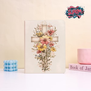 May include: A cream-colored journal featuring a floral cross design with pink and yellow flowers. The cross is centered on the cover, with the flowers arranged around it. A blue bubble candle and a pink mug are on the table.