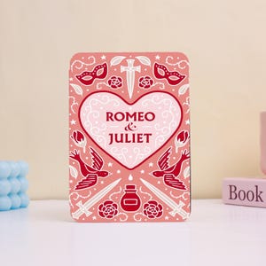May include: A rectangular, pink decorative item featuring the text "Romeo & Juliet" in a heart-shaped design. The design includes red illustrations of birds, swords, roses, and masks. The item is placed on a white surface.