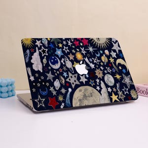 Peut inclure: Un ordinateur portable avec une coque bleu marine orn&eacute;e d'un motif c&eacute;leste. Le motif comprend diverses &eacute;toiles, lunes, plan&egrave;tes et soleils multicolores. L'ordinateur portable a un logo Apple blanc.