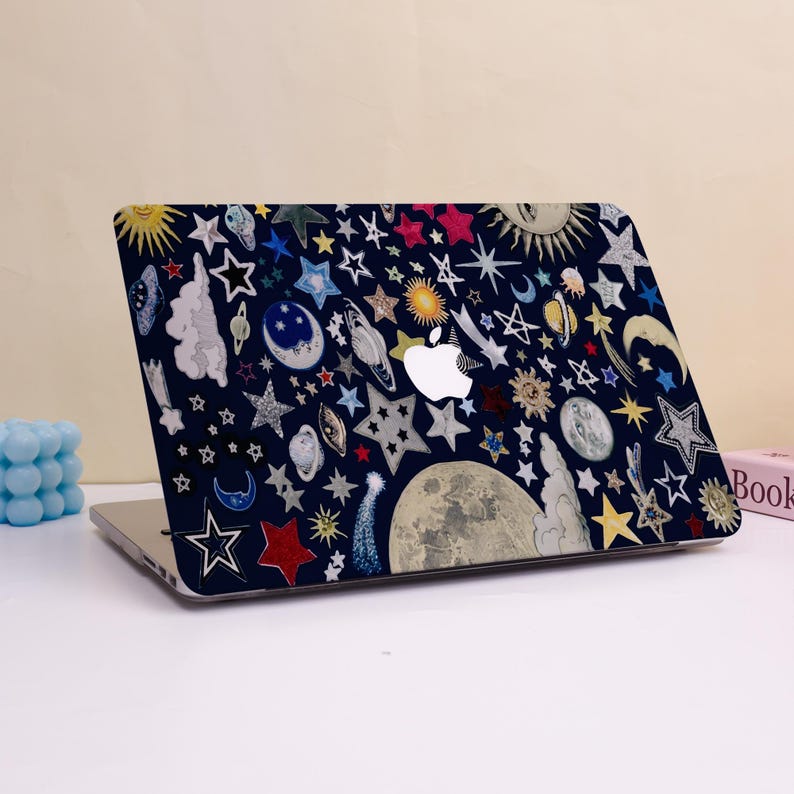 Peut inclure: Un ordinateur portable avec une coque bleu marine orn&eacute;e d'un motif c&eacute;leste. Le motif comprend diverses &eacute;toiles, lunes, plan&egrave;tes et soleils multicolores. L'ordinateur portable a un logo Apple blanc.