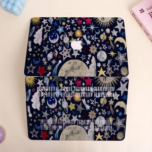 Peut inclure: Une coque d'ordinateur portable bleu fonc&eacute; avec un motif c&eacute;leste compos&eacute; d'&eacute;toiles, de lunes, de soleils et de plan&egrave;tes de diff&eacute;rentes couleurs. Le logo Apple est visible. Le motif est r&eacute;p&eacute;t&eacute; sur la moiti&eacute; inf&eacute;rieure de l'ordinateur portable.