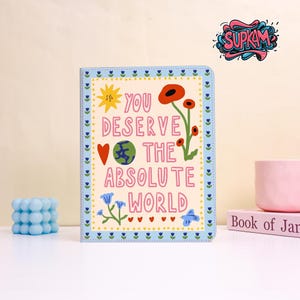 Könnte beinhalten: Ein farbenfrohes Notizbuch mit hellblauem Rand und den Worten "YOU DESERVE THE ABSOLUTE WORLD." Das Cover zeigt Illustrationen von Sonne, Blumen, einem Herzen, einem Globus und einem Schmetterling. Ein blauer Blasenwürfel und eine rosa Tasse sind ebenfalls zu sehen.