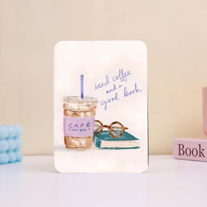 Puede incluir: Una impresión rectangular con una ilustración en acuarela de un café helado, un libro y gafas redondas. El texto "iced coffee and a good book" está escrito en guión azul. La taza de café tiene una etiqueta morada que dice "CAFE - new york".