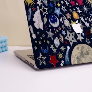 Peut inclure: Un ordinateur portable avec une coque bleu marine orn&eacute;e d'un motif c&eacute;leste. Le motif pr&eacute;sente diverses &eacute;toiles, plan&egrave;tes, lunes et soleils dans des tons de blanc, argent, rouge et jaune. Le logo Apple est visible.