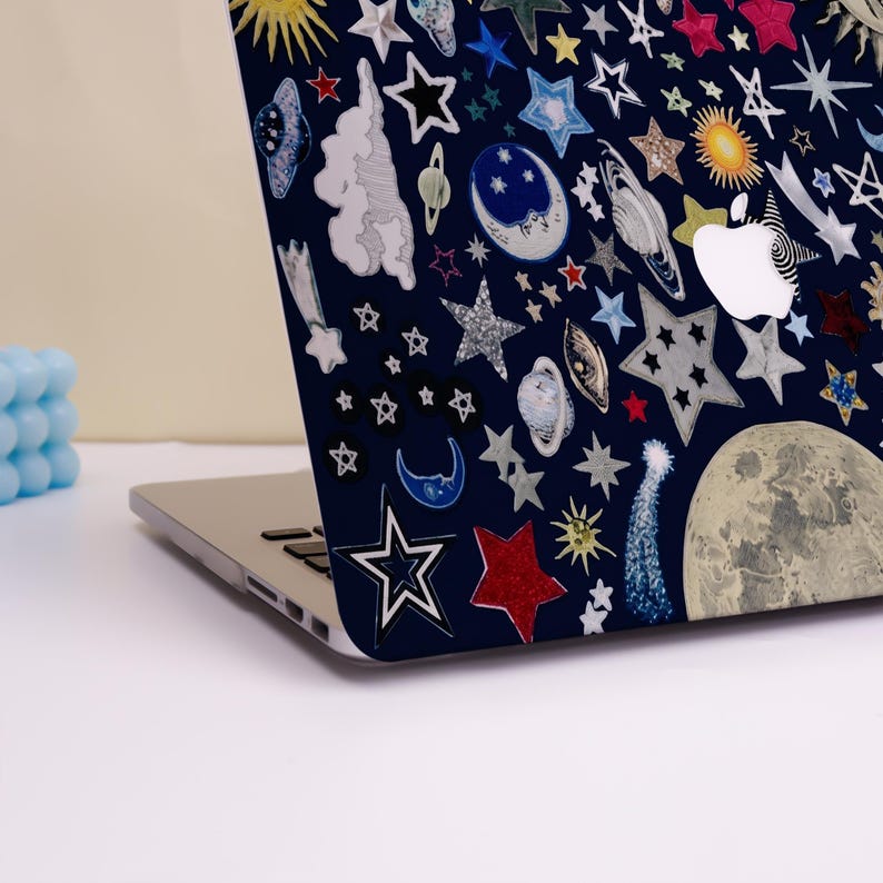 Peut inclure: Un ordinateur portable avec une coque bleu marine orn&eacute;e d'un motif c&eacute;leste. Le motif pr&eacute;sente diverses &eacute;toiles, plan&egrave;tes, lunes et soleils dans des tons de blanc, argent, rouge et jaune. Le logo Apple est visible.