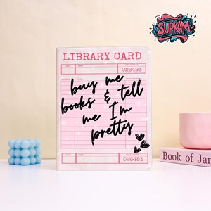 May include: A rectangular notebook with a light pink cover designed to look like a vintage library card. The text reads LIBRARY CARD and buy me books & tell me I'm pretty in black script. Three small black hearts are printed in the bottom right corner.