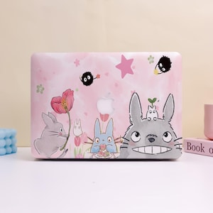 May include: Pink laptop case featuring a whimsical illustration of animated characters. The design includes a large gray rabbit with a wide grin, smaller blue and gray rabbits, and black, fluffy creatures. Pink flowers and a star accent the design.
