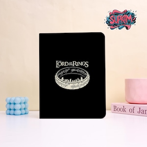 Puede incluir: Un cuaderno rectangular negro con un gráfico blanco del logotipo de El Señor de los Anillos y el Anillo Único con una silueta de personajes en su interior. La portada muestra la icónica inscripción del anillo en un patrón circular.