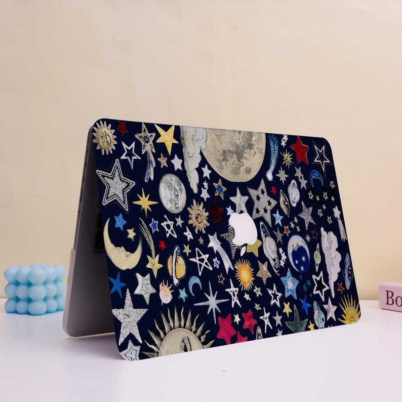 Peut inclure: Une coque d'ordinateur portable avec un fond bleu fonc&eacute; orn&eacute; d'illustrations c&eacute;lestes. Le motif comprend des &eacute;toiles, des lunes, des soleils et des plan&egrave;tes de diff&eacute;rentes couleurs et styles. Le logo Apple est visible.