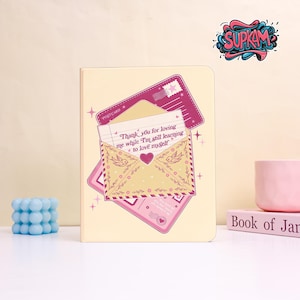 Puede incluir: Un cuaderno color crema con un diseño de sobre rosa y amarillo. El sobre tiene ilustraciones de corazón y pájaro, con el texto "Thank you for loving me while I'm still learning to love myself". Una postal rosa está detrás del sobre.