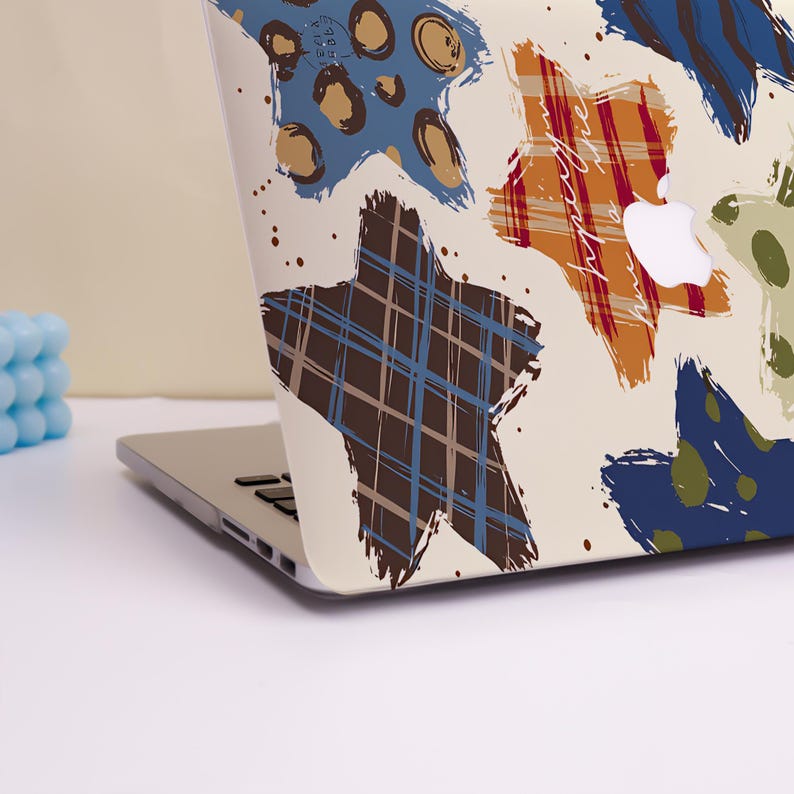 May include: Laptop case with a cream background and a colorful star pattern. The stars are in shades of blue, brown, orange, and green, with a plaid and dotted design. The Apple logo is visible.