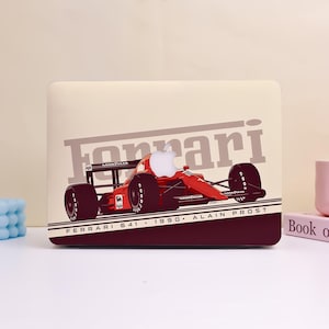 Puede incluir: Una funda para portátil con un diseño de coche de carreras Ferrari rojo. El diseño incluye el logotipo de Ferrari, el texto "FERRARI 641 • 1990 • ALAIN PROST" y el logotipo de Apple. El fondo es de color beige claro.