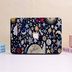 May include: A navy blue laptop case covered in colorful celestial illustrations. The design includes stars, moons, planets, and a sun, with a white Apple logo in the center. The case is decorated with various shapes and sizes of stars.