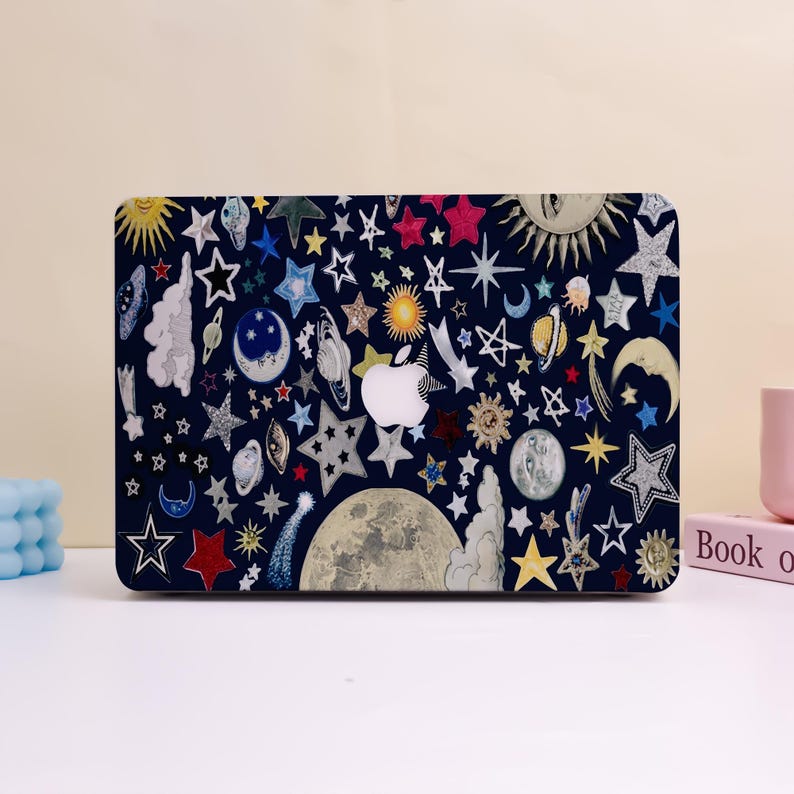 Peut inclure: Une coque d'ordinateur portable bleu marine orn&eacute;e d'illustrations c&eacute;lestes color&eacute;es. Le motif comprend des &eacute;toiles, des lunes, des plan&egrave;tes et un soleil, avec un logo Apple blanc au centre. La coque est d&eacute;cor&eacute;e d'&eacute;toiles de diff&eacute;rentes formes et tailles.