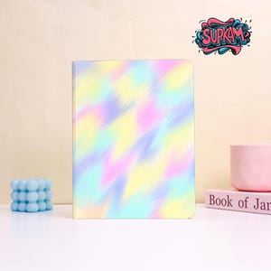 Puede incluir: Un diario de colores pastel con un diseño abstracto en tonos amarillos, rosas, azules y morados. El diario es rectangular y está en posición vertical. Una vela azul en forma de cubo y una taza rosa están a cada lado. El texto "SUPKAM" está en la esquina superior derecha.