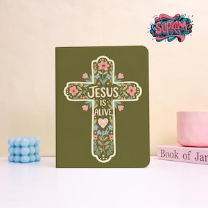 Peut inclure: Un journal vert olive avec un motif de croix florale et les mots "Jésus est vivant". La croix est ornée de fleurs roses et bleues et de feuilles vertes. Un cœur rose est au centre de la croix.