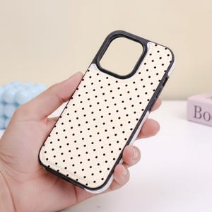 May include: A cream-colored phone case with a black polka dot pattern. The case has a black border around the camera and a white border around the edges. The case is designed to protect a phone.