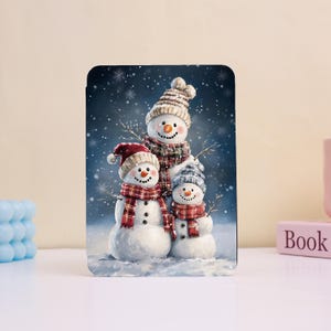 May include: A rectangular winter-themed image featuring three snowmen. Each snowman wears a hat and a scarf. The background is a dark blue with falling snowflakes. The snowmen have black dot eyes and button noses.