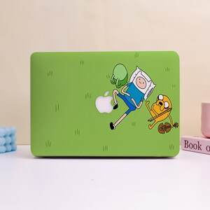 May include: A bright green laptop hard shell case featuring a graphic of Finn the Human lying down and Jake the Dog playing a brown violin. The design includes a white Apple logo cutout integrated into the scene, set against a grassy field background.
