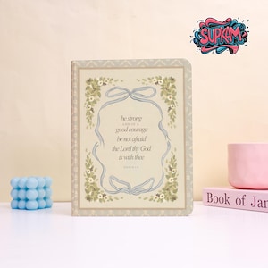May include: A decorative journal with a floral design and the text "be strong and of a good courage be not afraid the Lord thy God is with thee." The journal is surrounded by a light blue ribbon.