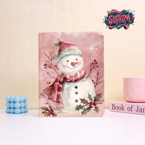 May include: A pink journal featuring a watercolor illustration of a snowman wearing a hat and scarf. The snowman has a carrot nose and is surrounded by snowflakes and holly. The journal has the text "SUPKAM" in the upper right corner.